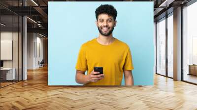 Young Moroccan man isolated on blue background using mobile phone Wall mural