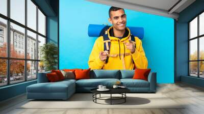 Young mountaineer man with a big backpack over isolated blue background points finger at you Wall mural