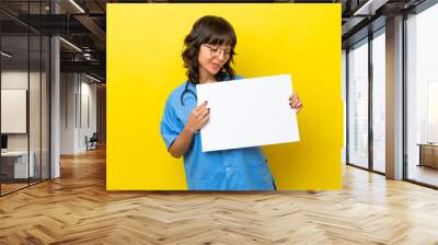 Young nurse doctor woman isolated on yellow background holding an empty placard with happy expression Wall mural