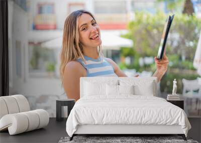 Young pretty woman holding a tablet at outdoors and pointing it Wall mural