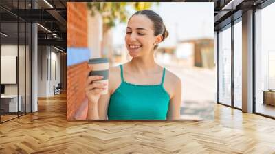 Young pretty woman holding a take away coffee at outdoors with happy expression Wall mural