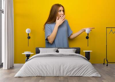 Young redhead girl over yellow wall background pointing finger to the side with a surprised face Wall mural