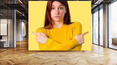 Young redhead girl with yellow sweater pointing to the laterals having doubts. Undecided person on isolated yellow background Wall mural