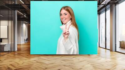 Young redhead woman isolated on blue background smiling and showing victory sign Wall mural