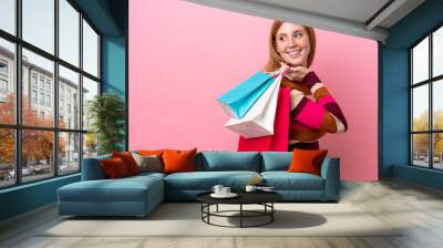 Young redhead woman isolated on pink background holding shopping bags and looking back Wall mural