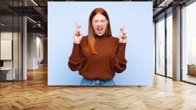 Young redhead woman over isolated blue background with fingers crossing Wall mural