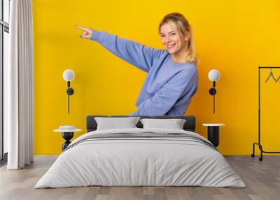 Young Russian woman isolated on yellow background pointing finger to the side and presenting a product Wall mural