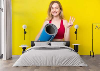 Young sport caucasian woman going to yoga classes while holding a mat isolated on yellow background happy and counting three with fingers Wall mural