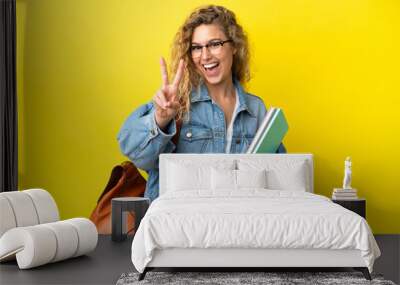 Young student caucasian woman isolated on yellow background smiling and showing victory sign Wall mural