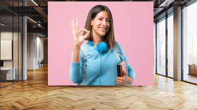 Young student girl with blue sweater and headphones showing an ok sign with fingers. Face of happiness and satisfaction on pink background Wall mural