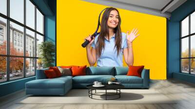 Young tennis player woman over isolated yellow wall saluting with hand with happy expression Wall mural