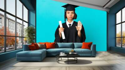 Young university graduate girl over isolated blue background making stop gesture Wall mural