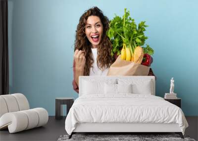 Young woman holding a grocery shopping bag isolated on blue background celebrating a victory in winner position Wall mural