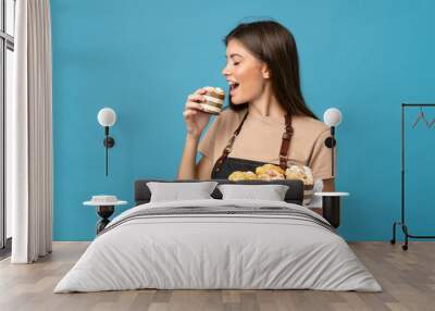 Young woman over isolated blue background holding mini cakes and eating it Wall mural