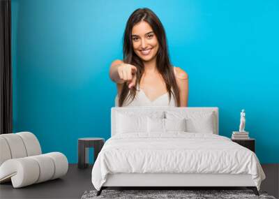 Young woman over isolated blue background points finger at you while smiling Wall mural