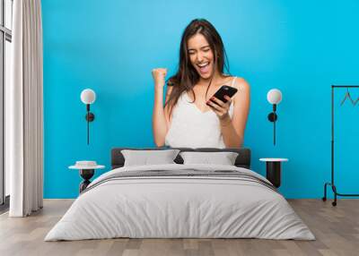 young woman over isolated blue background with phone in victory position Wall mural