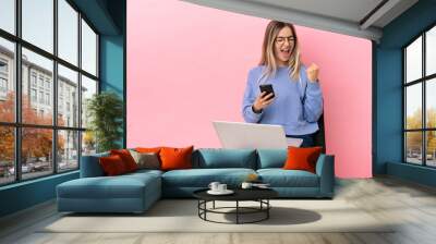 Young woman sitting on a chair with laptop over isolated pink background with phone in victory position Wall mural