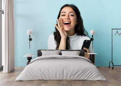 Young woman using mobile phone isolated on blue background shouting with mouth wide open Wall mural