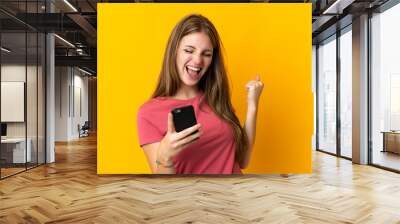 Young woman using mobile phone isolated on yellow background celebrating a victory Wall mural