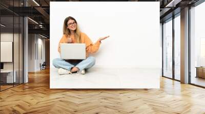 Young woman with a laptop sitting on the floor holding copyspace imaginary on the palm to insert an ad and with thumbs up Wall mural