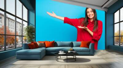 Young woman with red sweater over isolated blue background extending hands to the side for inviting to come Wall mural