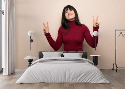 Young woman with red turtleneck smiling and showing victory sign with both hands Wall mural