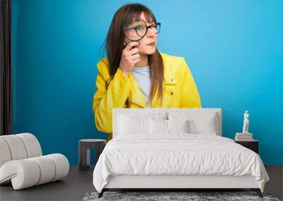 Young woman with yellow jacket on blue background taking a magnifying glass and looking through it Wall mural