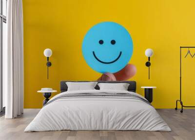 Happy face icon held on yellow background. A hand holding a blue smiley face icon against a vibrant yellow backdrop. This cheerful image conveys positivity and optimism. Wall mural