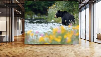 A black bear stands in a rushing river, surrounded by yellow and pink wildflowers. Wall mural