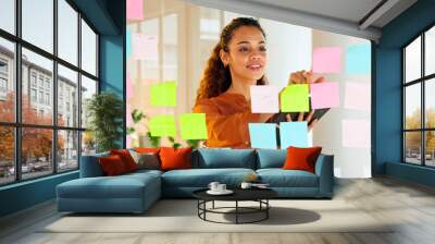 Happy female designer planning ideas on a glass wall with colorful sticky notes inside a creative and modern office. Busy woman enjoying her job while brainstorming projects and managing projects Wall mural