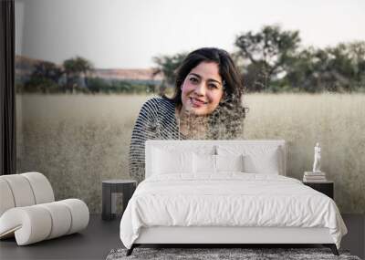 Young adult smiling Mexican woman crouched in a dry grassy meadow looking at the camera Wall mural