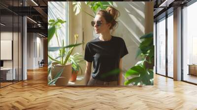 Fashionable woman in black outfit and sunglasses by a window. Indoor chic style portrait with palm plant backdrop. Urban elegance and fashion concept for design and print. Side view with natural light Wall mural