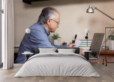 Senior man learn to use computer. Old man in glass and blue shirt Enters a password from a piece of paper. Concept using a laptop computer for online studying at home office Wall mural