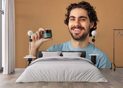 Facial skin care. Young brunette curly man holding skin care cream and looking at camera against beige studio background. Concept of male facial care routine, cosmetic product, anti-aging. Wall mural