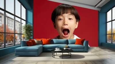 Little Asian oy, child playfully yawning or shouting with mouth wide open, showing energy against bold red studio background. Concept of facial-expressions, positive and negative emotions. Ad Wall mural