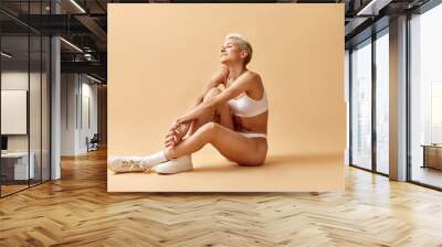 Portrait of beautiful young woman with short blonde hair sitting on floor in underwear and sneakers, posing against yellow studio background Wall mural