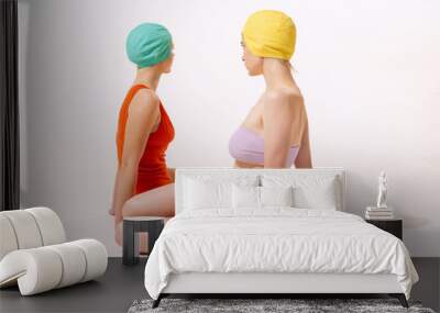 Portrait of two young women in swimming suit and cap sitting, posing isolated over grey studio background Wall mural