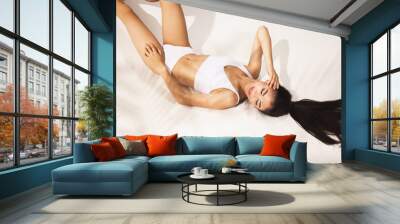 Portrait of young beautiful tanned woman in white underwear lying on floor over white background. Wall mural