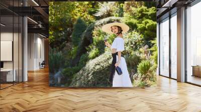 Young woman with wide-brimmed hat, holding glass of red wine, stands in verdant garden, embodying tradition of Mediterranean winemaking. Concept of end of summer, harvesting, travelling, culture. Wall mural