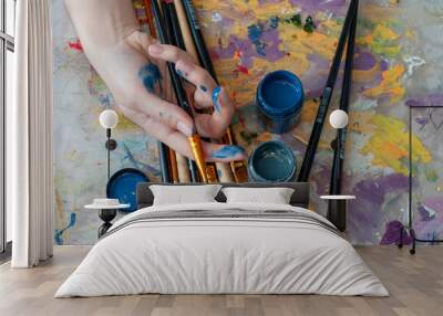 The artist's close-up hand holds a brush against the background of a palette, brushes and paints, is engaged in art, creativity, hobbies, anti-stress or art therapy. Wall mural