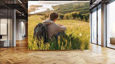 Tourist with backpack sitting on top of hill in grass field and enjoying beautiful landscape view. Rear view of teenage boy hiker resting in nature. Active lifestyle. Concept of local travel Wall mural