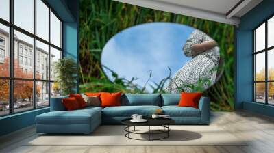 An oval mirror lies on the grass and reflects the waist of a woman in a beautiful dress. A fashionable image close to the nature concept. Blue sky reflected in the mirror. Trendy image close-up. Wall mural