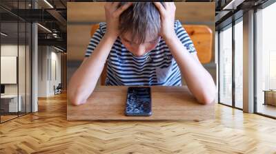 Portrait of scared kid girl with broken mobile phone on table. Sad child broke screen of smartphone. Teen boy in desperation is holding his head and looking at phone with cracked screen at day time Wall mural