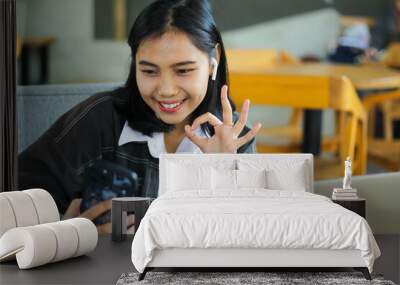 carefree and attractive asian woman sitting in cafe using smartphone to do video call and showing okay symbol with laptop on desk Wall mural