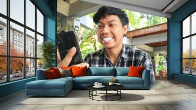 young asian man smilling with white tooth while looking at black mobile phone feeling happy and excited gesture. cheerful young asian man wearing flannel shirts Wall mural