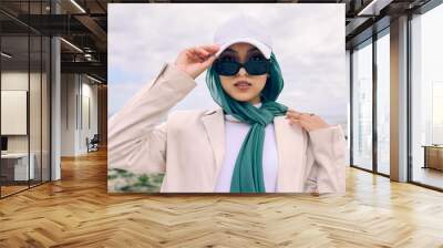 Portrait, fashion or sunglasses with a saudi woman outdoor in a cap and scarf for contemporary style. Islam, face and hijab with a trendy young muslim or arab person posing outside in modern clothes Wall mural