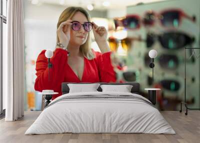 A woman in a red dress tries on glasses in a store. Wall mural