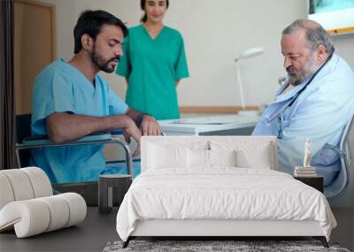 Asian senior male doctor treating knee pain to an Indian male patient sitting in a wheelchair in the treatment room at the hospital Wall mural