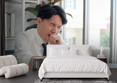 Exhausted young asian worker yawning at workplace. Business man using laptop for working feeling sleepy and yawning tired from overwork job Wall mural