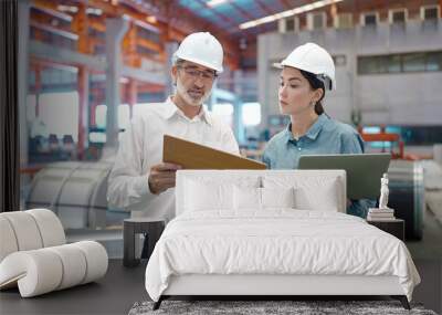 Two engineer manager leader and woman assistant holding laptop wearing helmet talking and checking production standing in workplace area at manufacturing factory Wall mural